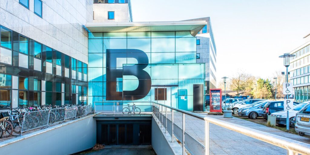 Glazen gevel met grote zwarte letter B bij gebouw aan het John M. Keynesplein, met fietsenstalling en parkeerplaatsen op de voorgrond.